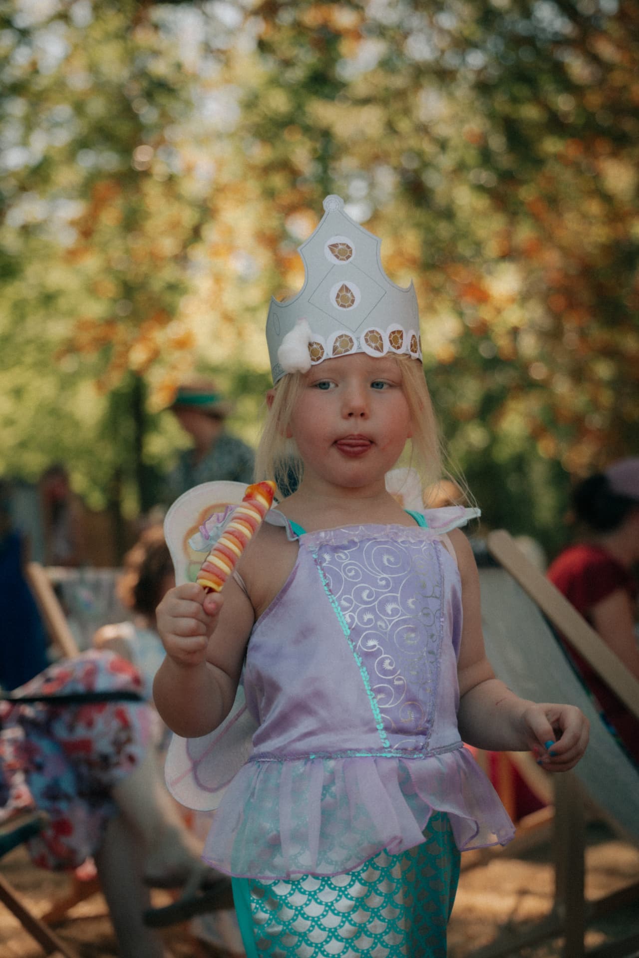 Een kind in een feeënkostuum met een papieren kroon en een lolly in de hand.